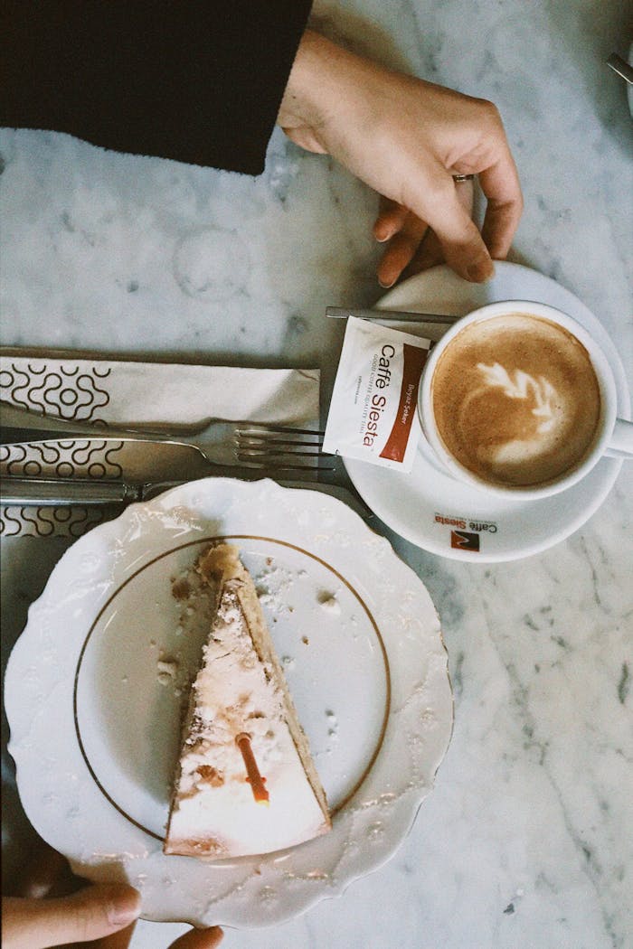 Top view of hands holding a cappuccino and cake slice at a café table, perfect for lifestyle and food themes.